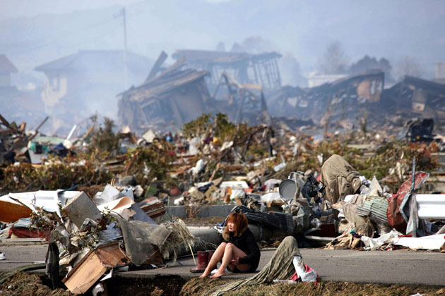 Женщина плачет, сидя на дороге, на фоне разрушенного города Натори.